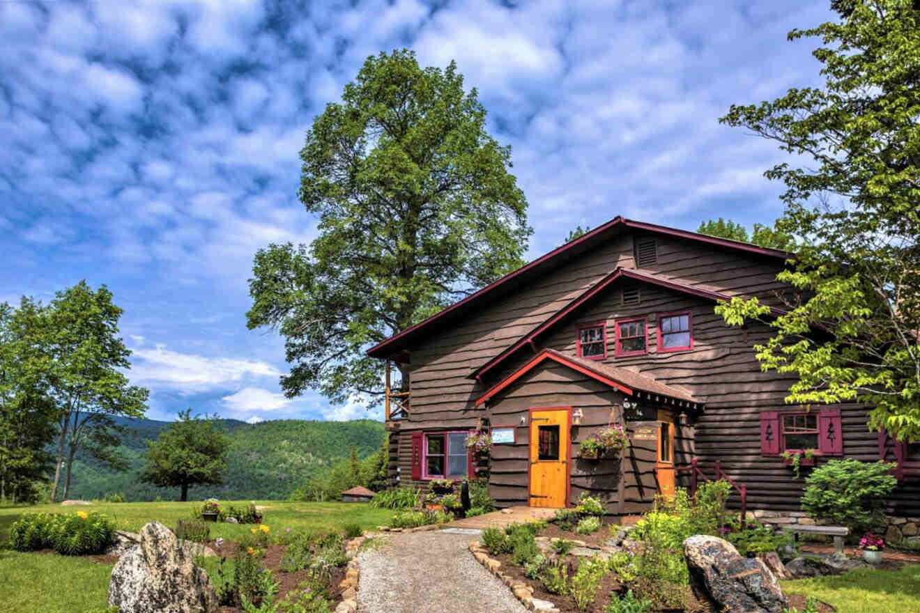 A rustic wooden cabin with red trim sits amidst green trees and a cloudy blue sky, with a mountain view in the background. A stone pathway leads to the entrance.