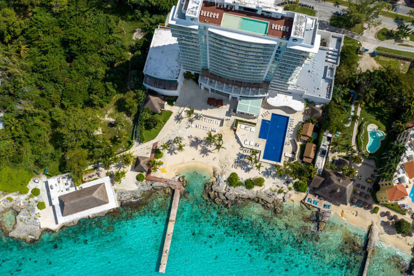 Aerial view of a beachfront hotel with a pool, surrounded by lush greenery and turquoise water, featuring a docking pier and sandy beach area.