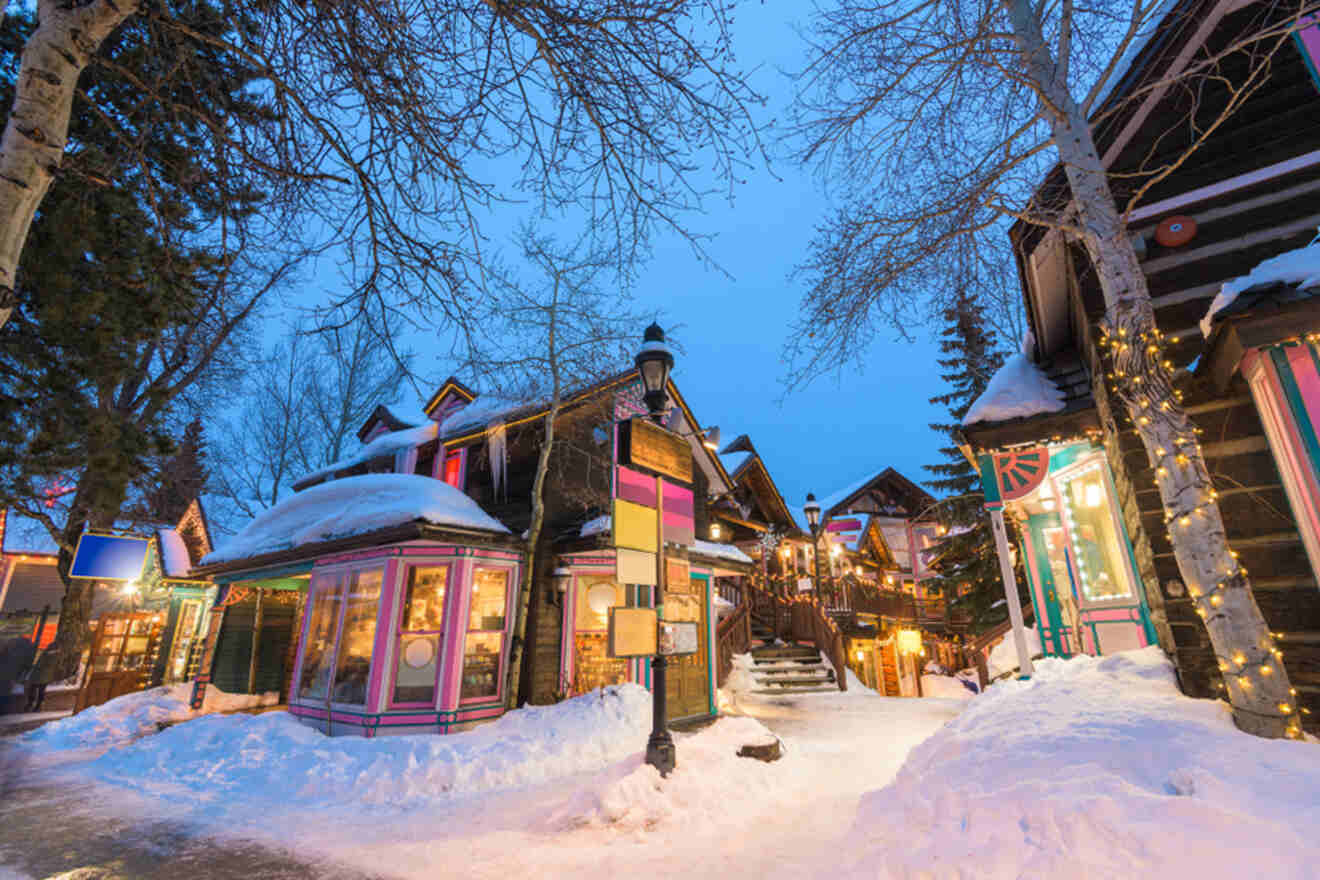 A snowy village scene at dusk, featuring colorful, illuminated shops and bare trees under a blue sky.