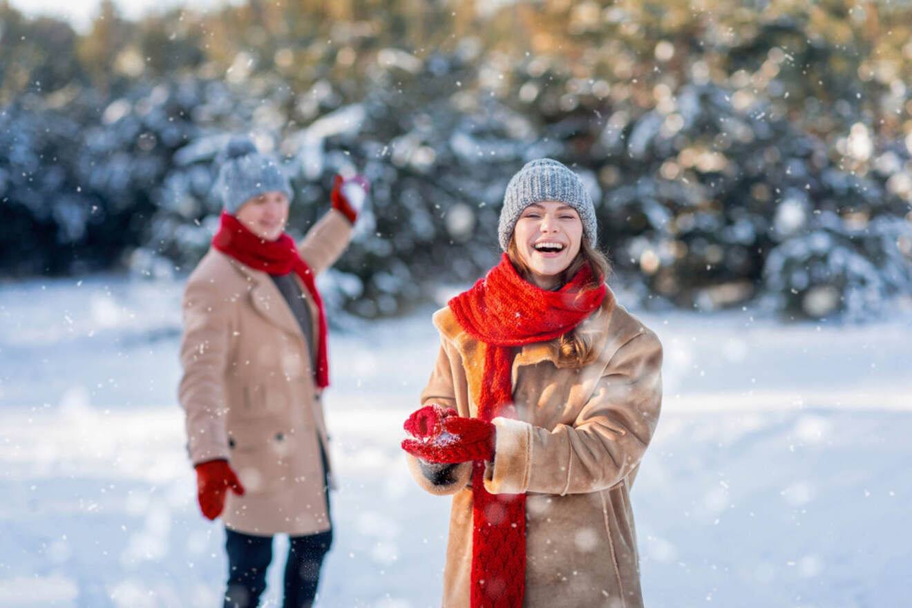 Two people in winter clothing, with red scarves and gloves, enjoying the snow in a snowy outdoor setting. One is holding a snowball and laughing, while the other is in the background.