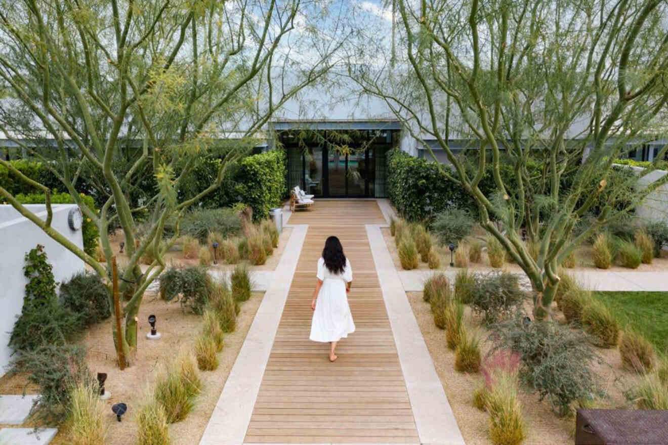 A person in a white dress walks down a wooden path surrounded by greenery, leading to a building entrance.