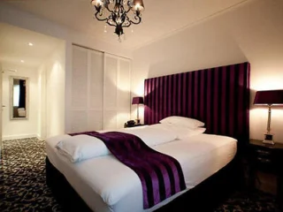A bedroom with a large bed featuring a purple striped headboard and matching throw. A chandelier hangs from the ceiling, and two bedside lamps are on either side of the bed.