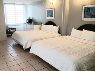 Two double beds in a hotel room with tiled floor, a small kitchenette in the corner, two framed pictures on the wall, and a window with blinds.