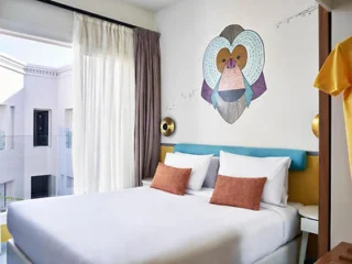 Hotel room with a neatly made bed, two orange pillows, large windows, and wall art of a stylized monkey face above the headboard.