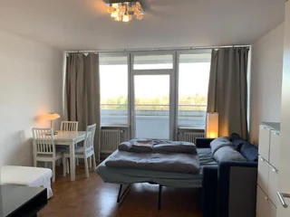 Studio apartment with a pull-out sofa bed, small dining table with chairs, and large windows with curtains. Ceiling light fixture and hardwood flooring.
