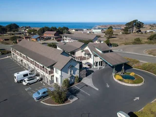 Aerial view of a motel with a parking area, surrounded by trees and buildings, near the coastline.