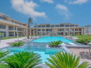 Swimming pool flanked by lounge chairs and palm plants, in front of a multi-story resort building under a blue sky.