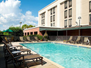 Outdoor hotel pool surrounded by lounge chairs, with a multi-story building in the background under a partly cloudy sky.