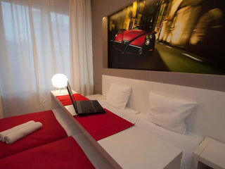 A modern bedroom with two single beds, red accents, a laptop on a desk, and a car-themed picture above the beds.