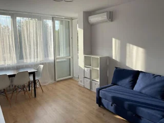 A bright, minimalistic room with a blue sofa, white shelving, a dining table with chairs, and a window letting in sunlight.