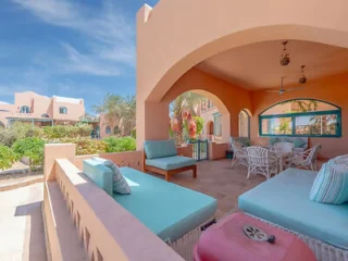 Patio with blue cushioned seating, dining area, and shaded arches. Desert-style architecture with pinkish walls and palm trees in the background under a clear sky.