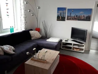 Living room with a dark sectional sofa, a wooden coffee table with white flowers, a TV on a stand, and cityscape photos on the wall. A red rug and tall lamp add accents.