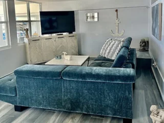 Modern living room with a dark teal sectional sofa, white coffee table, and a large TV. Decor includes a wall-mounted anchor and a small statue on the floor. Grey flooring and lots of light.