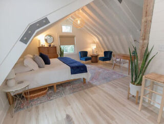 A cozy attic bedroom with a bed, dresser, two blue chairs, a desk, and potted plants. The space features slanted ceilings and wooden flooring, with soft natural light filtering in.