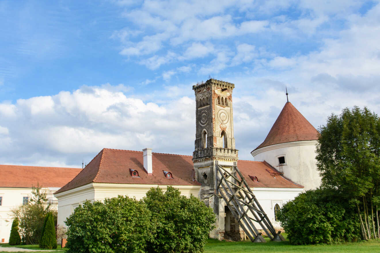 Historic building with a tall, ornate tower and red-tiled roofs, surrounded by greenery under a partly cloudy sky.