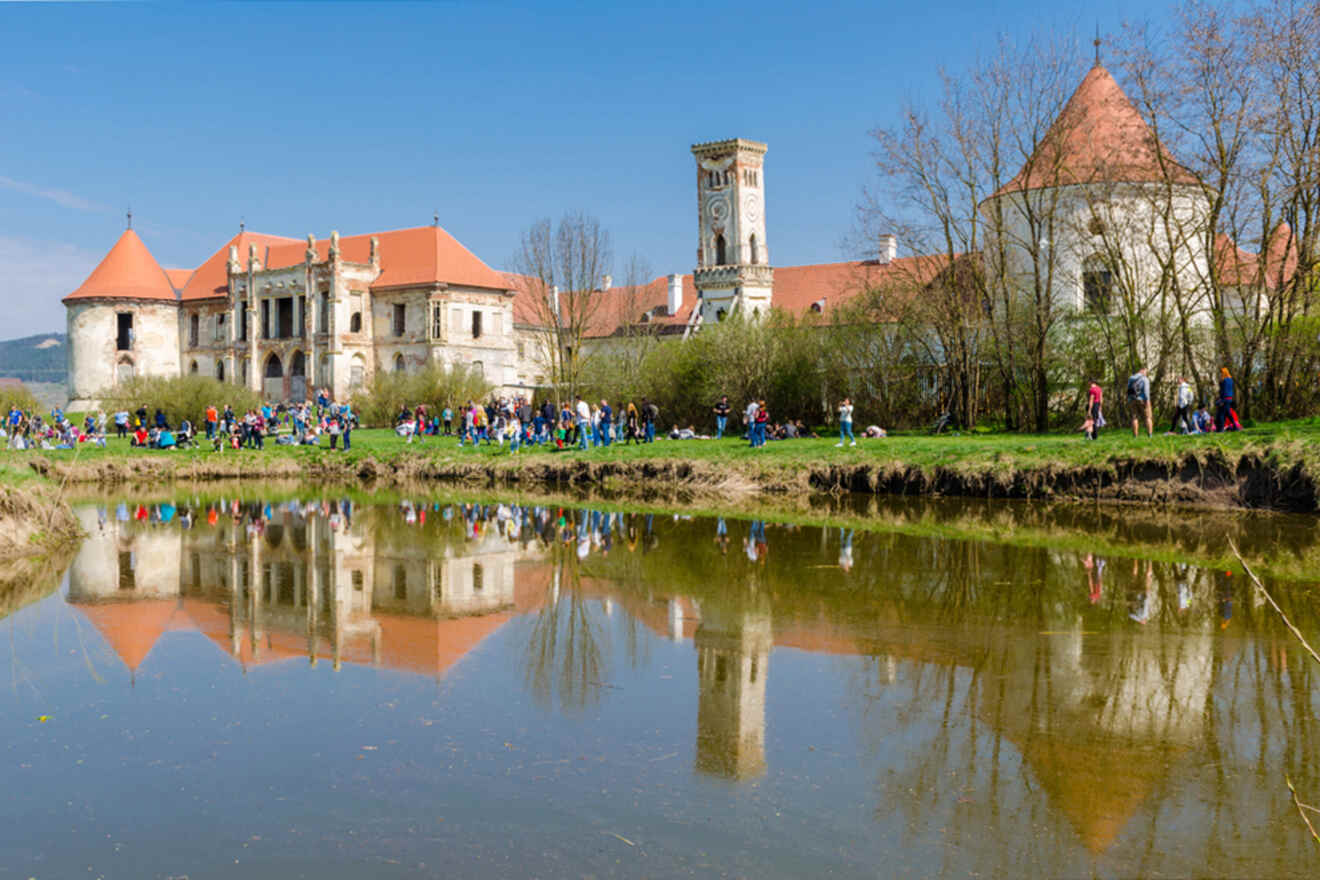 A historic castle with red roofs is reflected in a calm pond. A crowd of people is gathered along the water's edge, and trees border the scene under a clear blue sky.