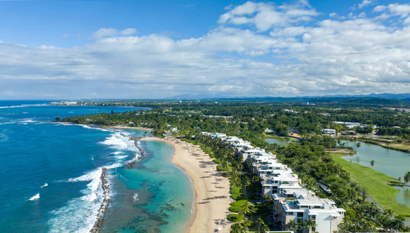 Aerial view of a coastline with a sandy beach, blue ocean, and adjacent greenery; buildings line the shore under a partly cloudy sky.