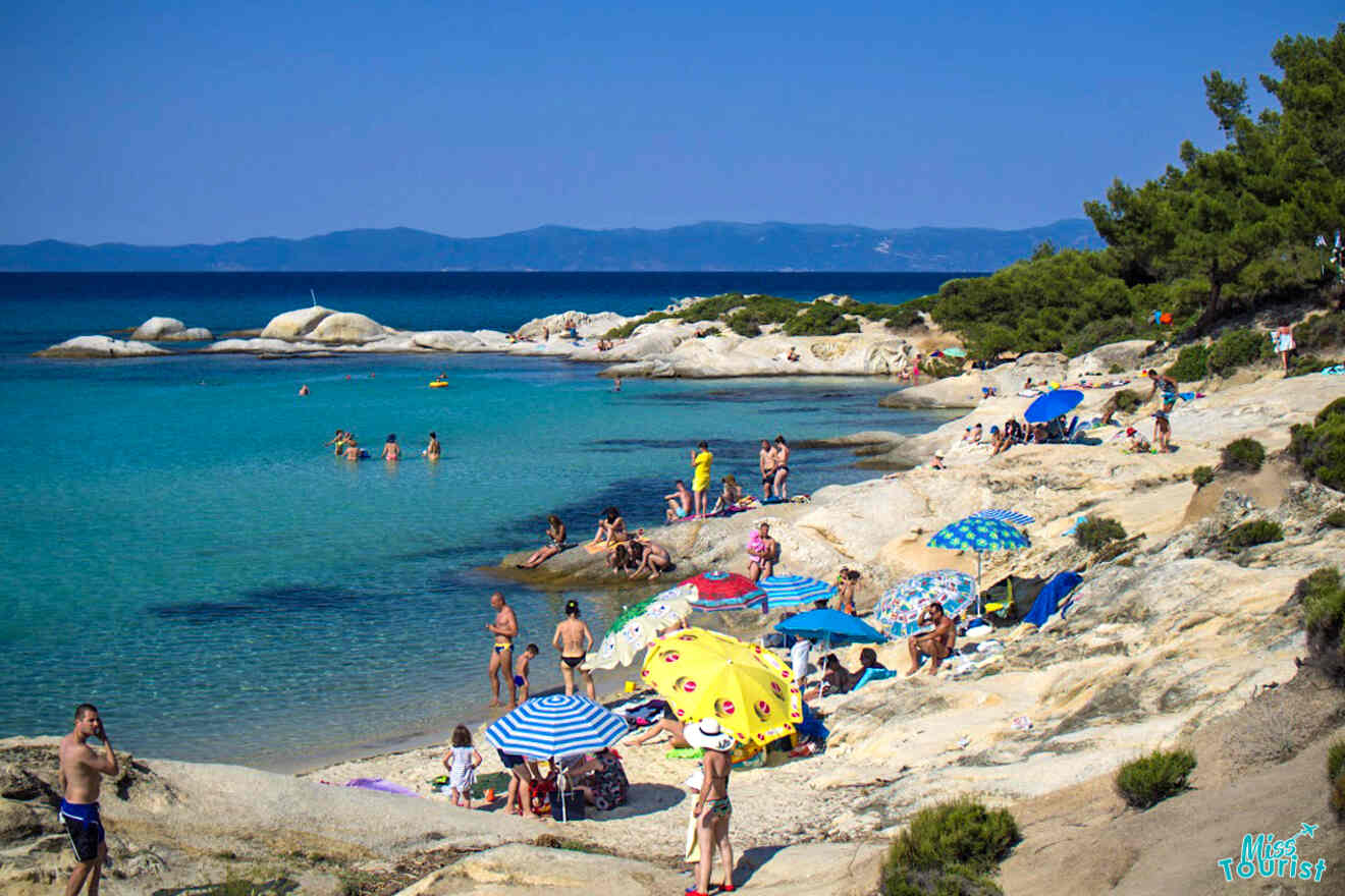 Beachgoers relax under colorful umbrellas on a rocky shoreline with clear blue water and distant hills in the background.