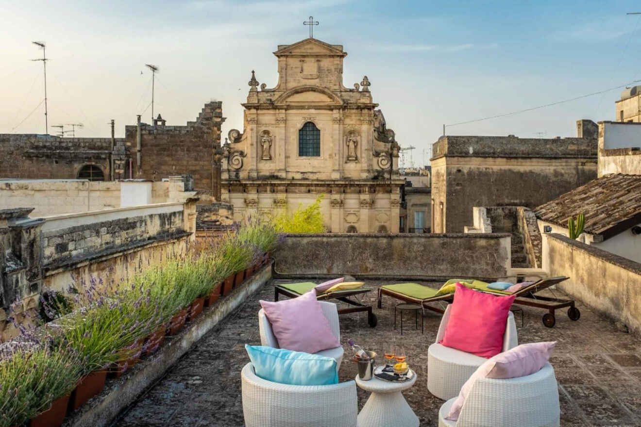 Rooftop terrace with chairs, colorful cushions, drinks, and loungers, overlooking an old stone building with a cross on top.