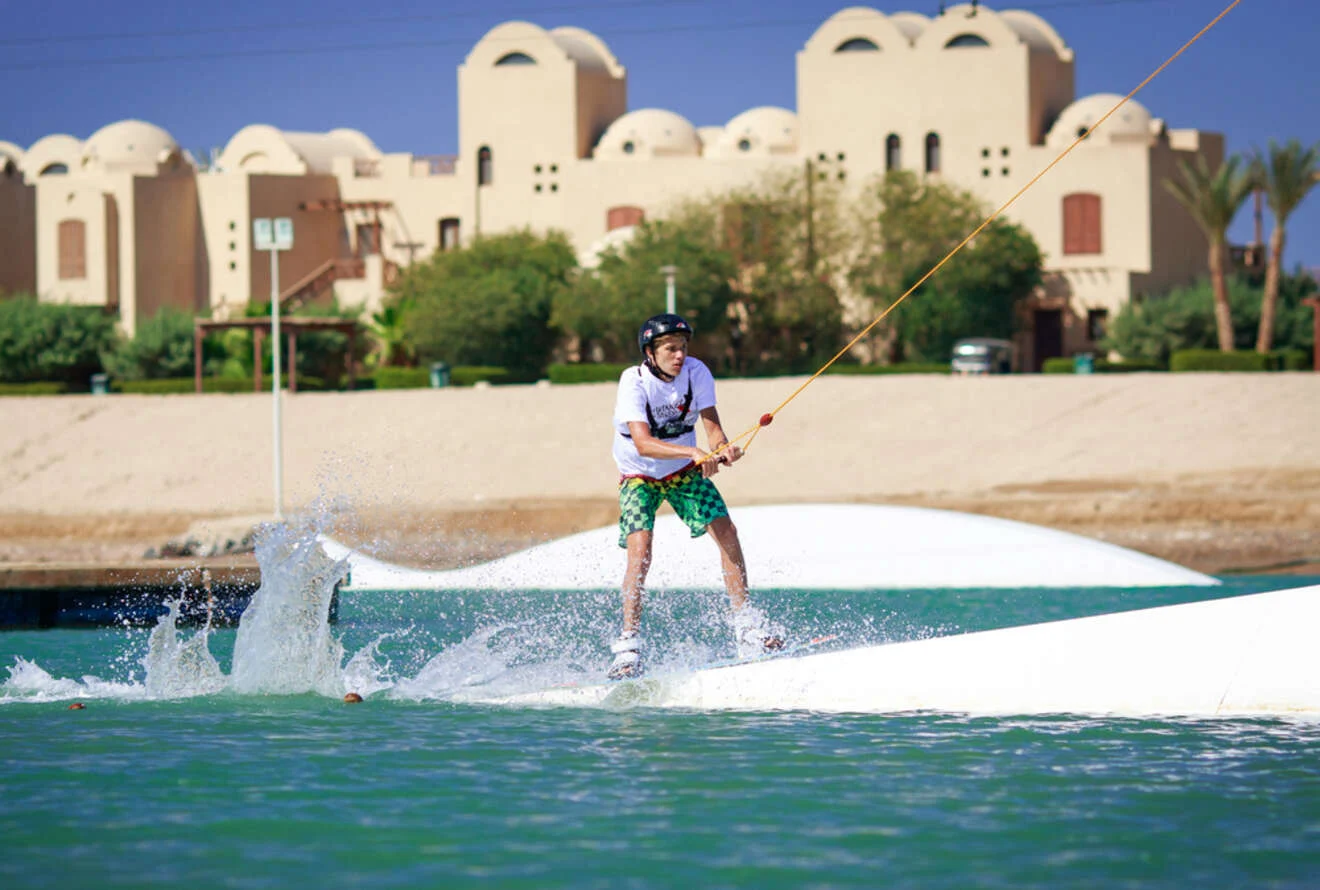 A person wakeboarding on a cable park with beige buildings in the background.