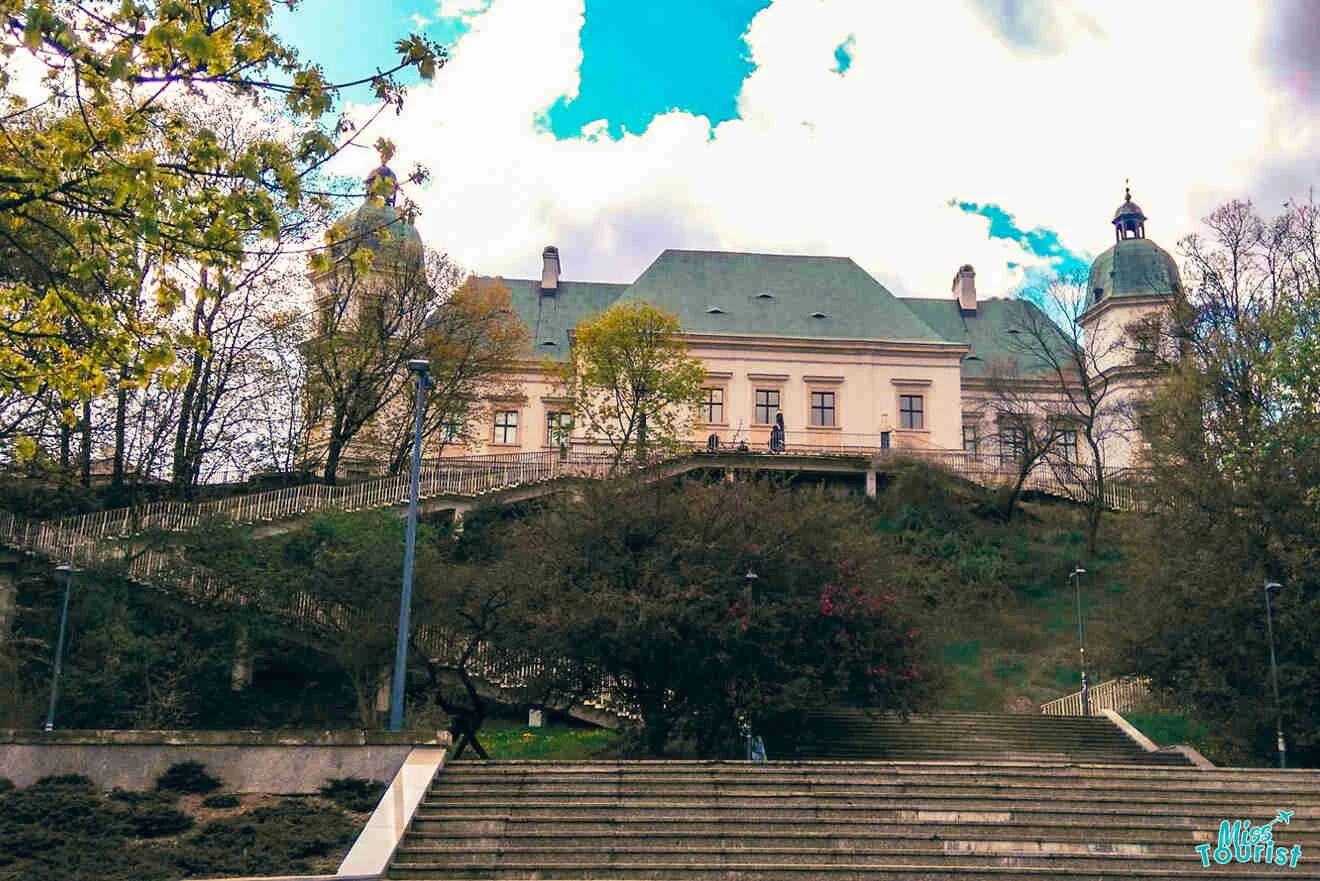 Historic building with towers and a steep staircase surrounded by trees, set against a partly cloudy sky.