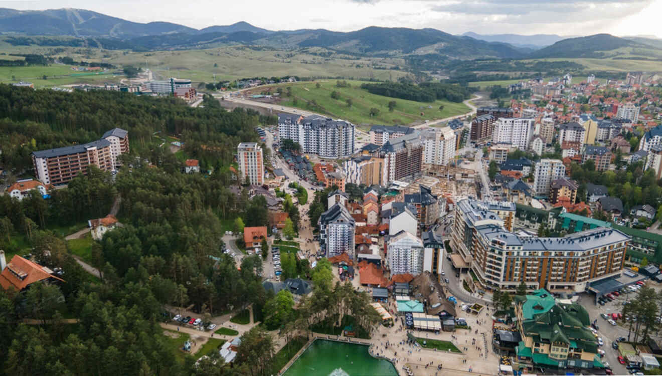 Aerial view of a small town surrounded by hills, featuring numerous buildings, roads, and a green pond in the foreground.