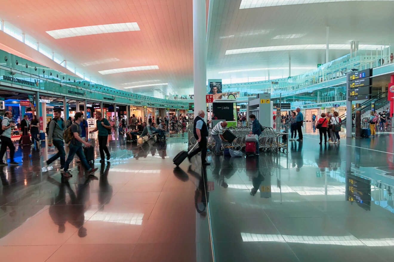 A busy airport terminal with people walking and sitting. Large windows and bright lighting illuminate the space. Various signs and shops are visible in the background.