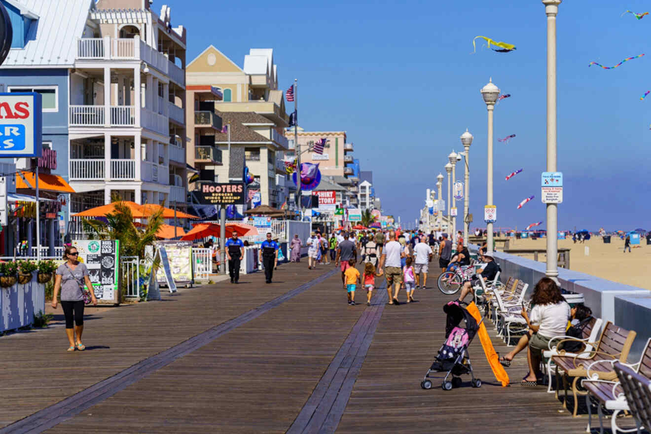 People walking and relaxing on a bustling boardwalk by the beach, with buildings and shops lining one side and beachgoers in the distance.