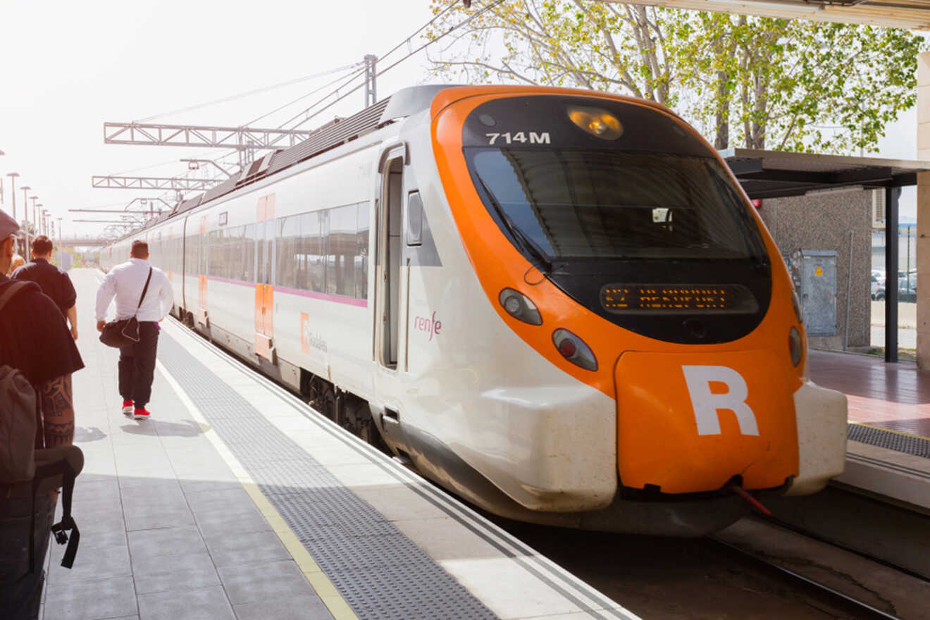 A white and orange commuter train is stopped at a sunny platform with passengers boarding and trees in the background.