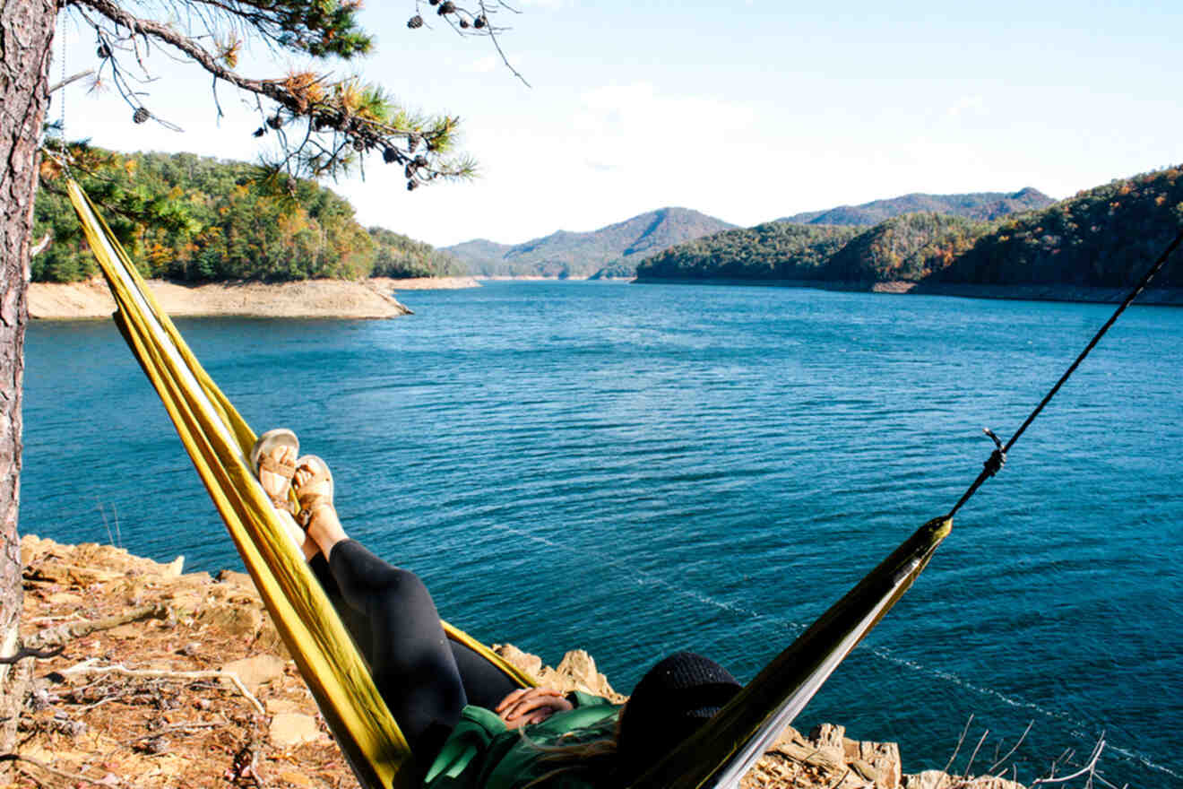 Person relaxing in a hammock by a lakeside, overlooking calm water and tree-covered hills under a clear sky.