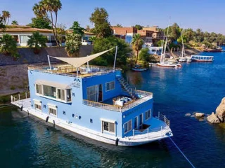 A blue houseboat with a rooftop seating area is moored on a river, surrounded by trees and residential buildings.