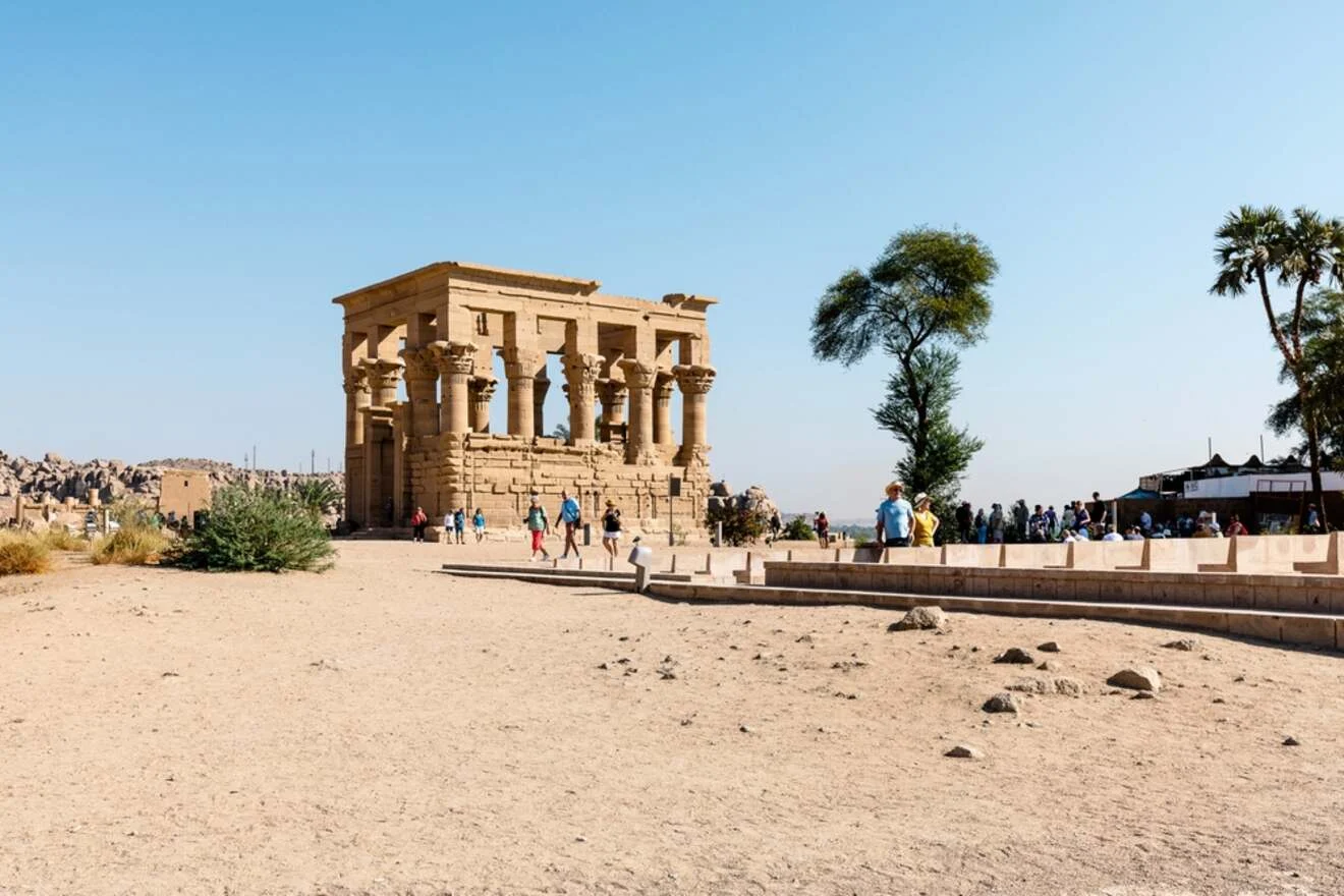 Ancient stone structure with columns in a desert landscape, surrounded by tourists and palm trees under a clear blue sky.