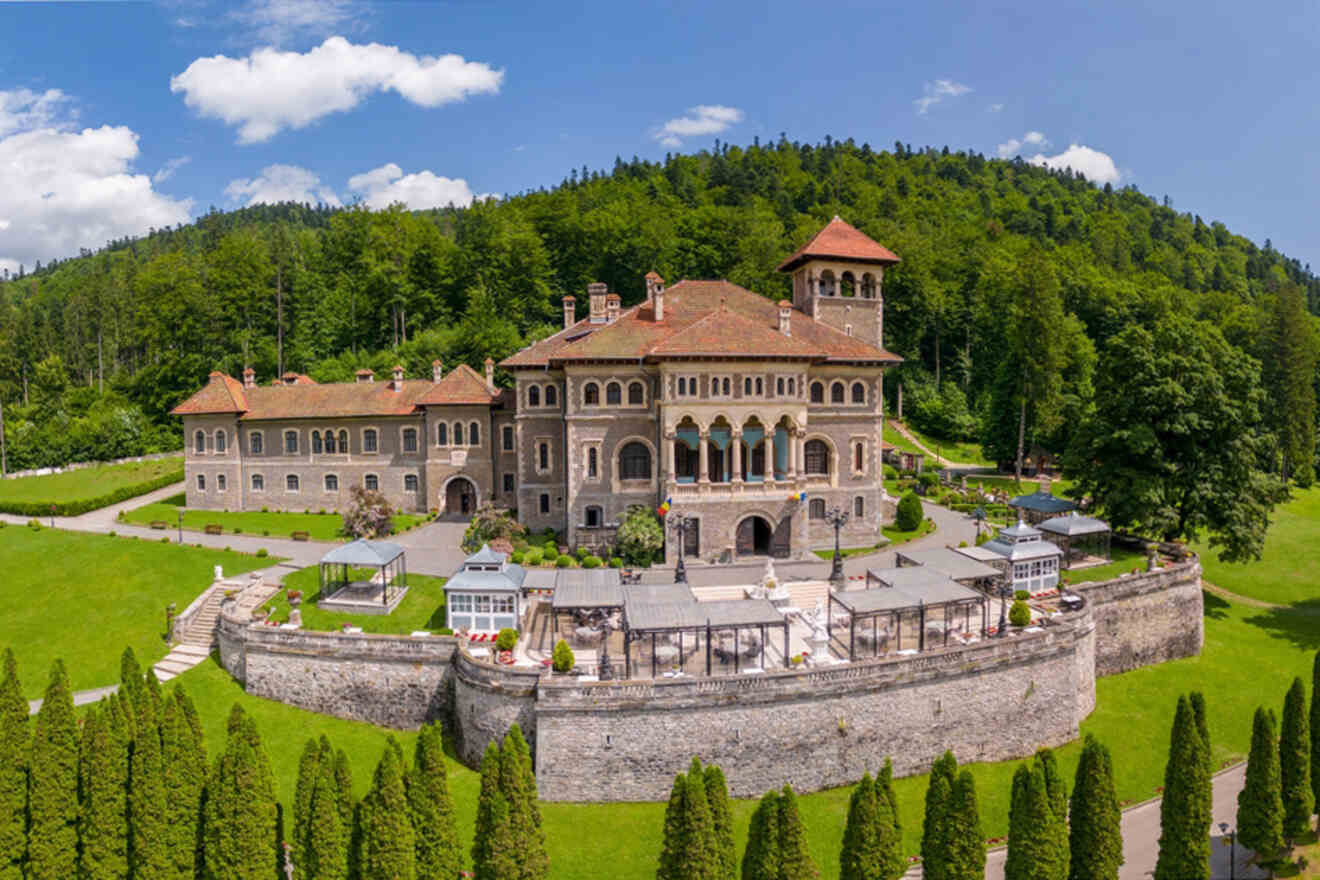 A large, historic castle with multiple towers and terraces, surrounded by a stone wall and lush green trees, under a partly cloudy sky.