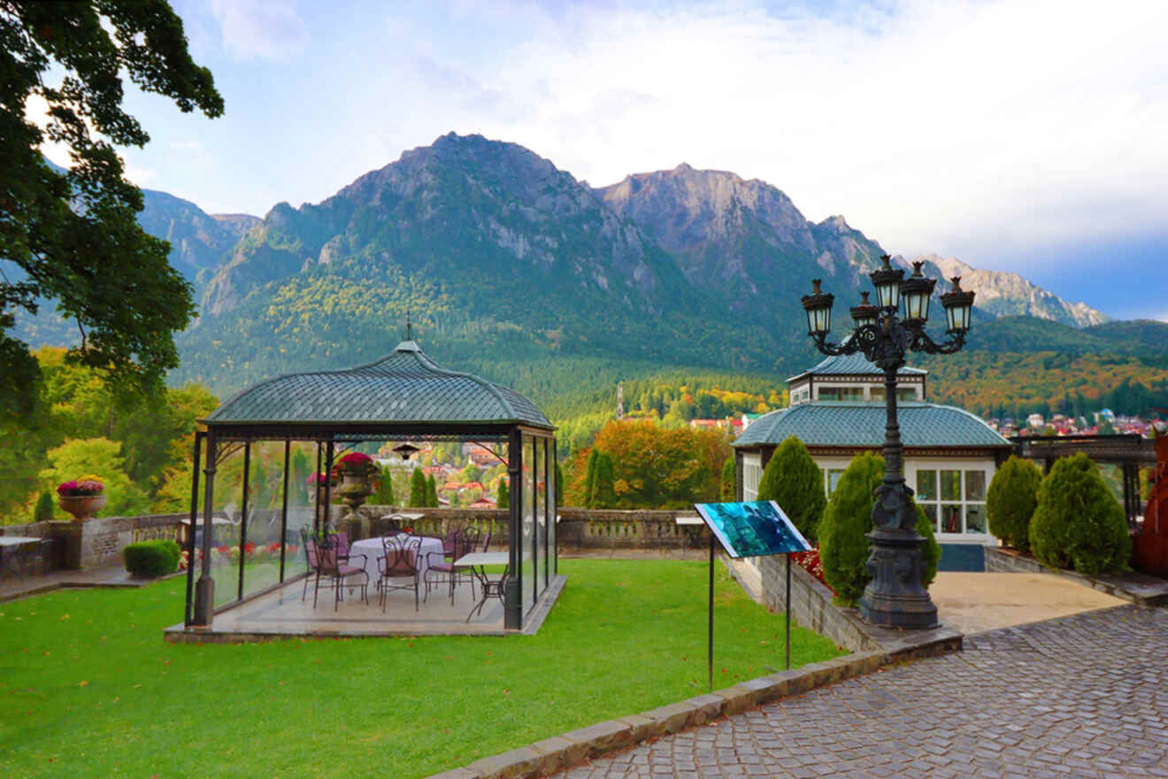 A glass gazebo on a grassy lawn with outdoor furniture inside. A paved path leads to a lamppost. In the background, there are mountains and trees under a partly cloudy sky.