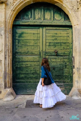 The writer of the post in a white dress and blue jacket stands in front of a large, weathered green door.