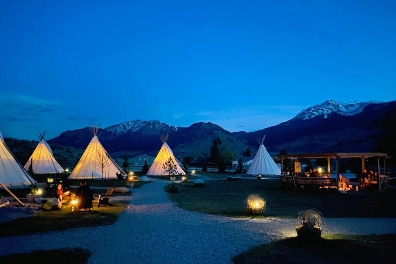 A nighttime scene of a campsite with lit tipis and small fires against a backdrop of silhouetted mountains and a clear sky.