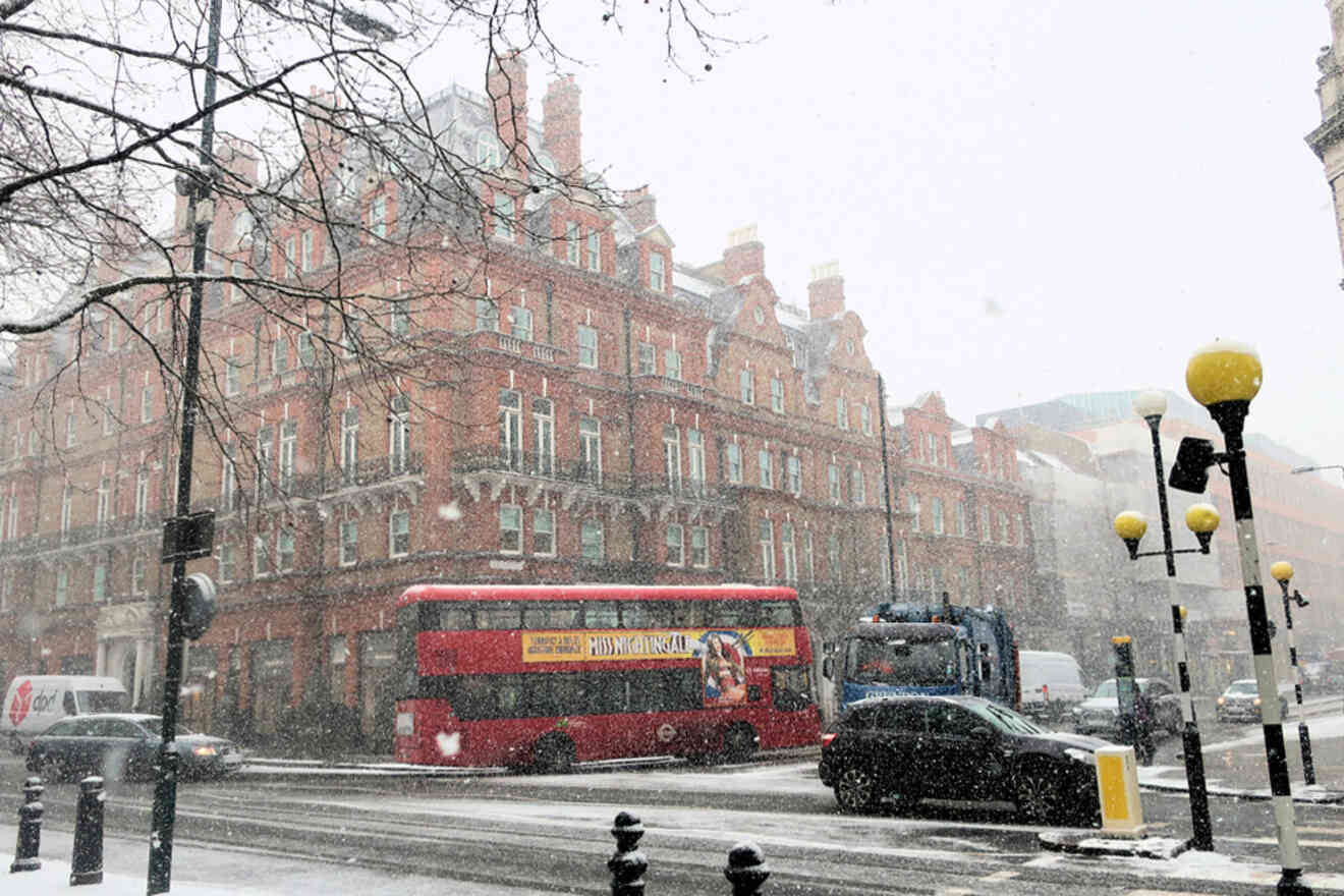 A red double-decker bus and other vehicles navigate a snowy street in front of a historical red brick building.