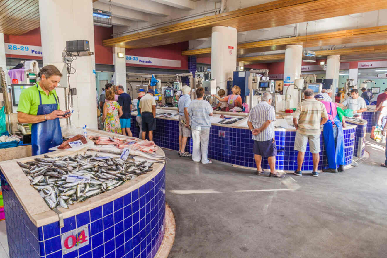 People browse and shop at a bustling indoor seafood market in Lagos, Portugal, with various fish on display in blue-tiled stalls.