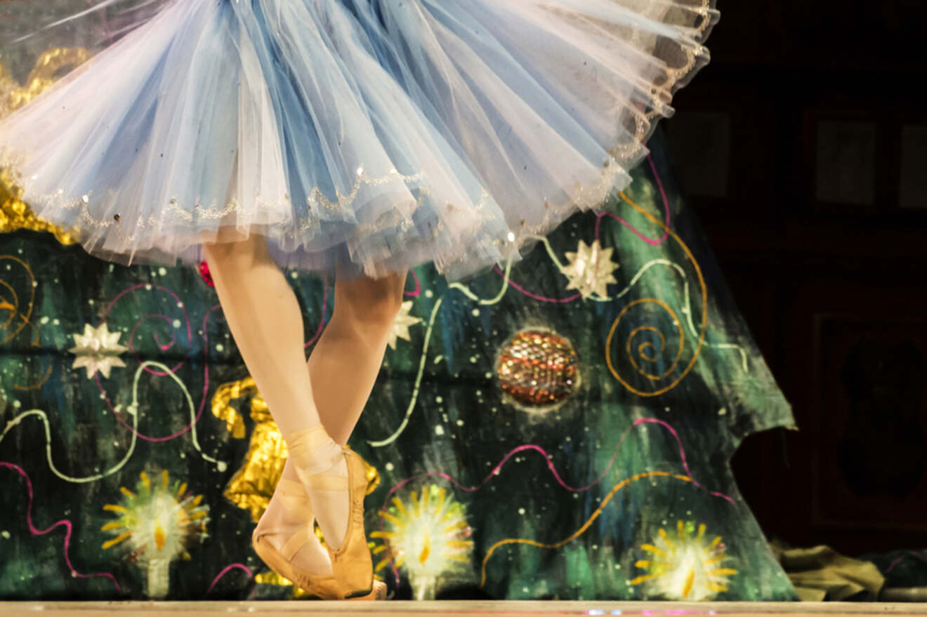 Ballet dancer wearing a blue tutu on pointe in front of a colorful stage backdrop.