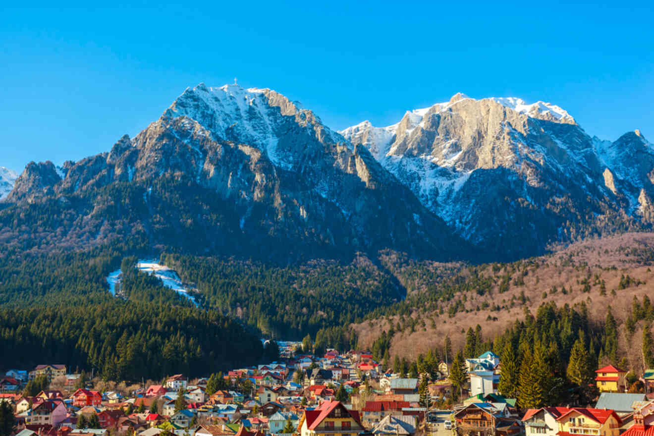 A colorful town with red-roofed buildings sits at the base of snow-capped mountains under a clear blue sky.