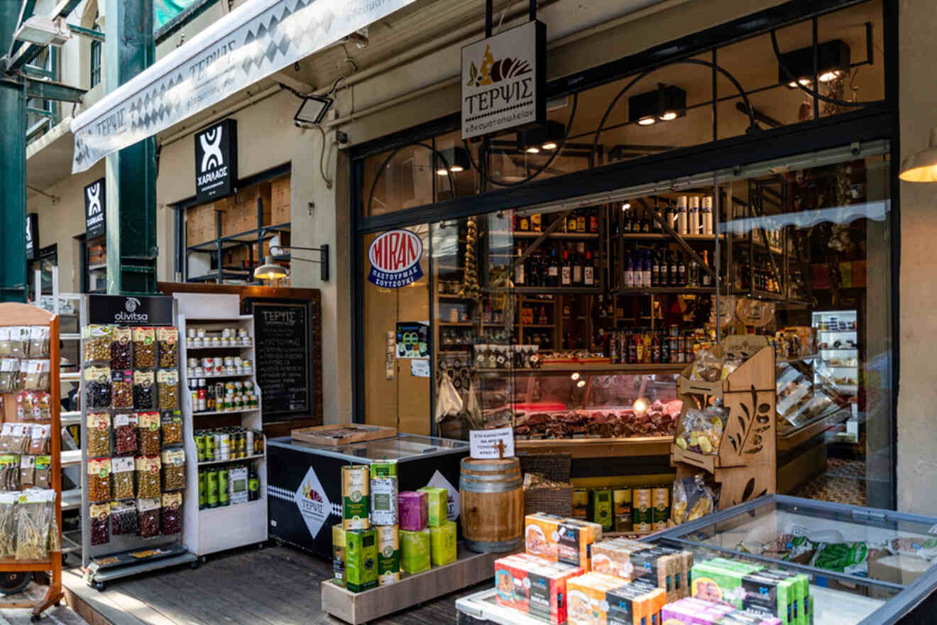 Street view of a delicatessen with a variety of packaged goods and a glass display of meats inside. The storefront features racks of products and a colorful, inviting atmosphere.