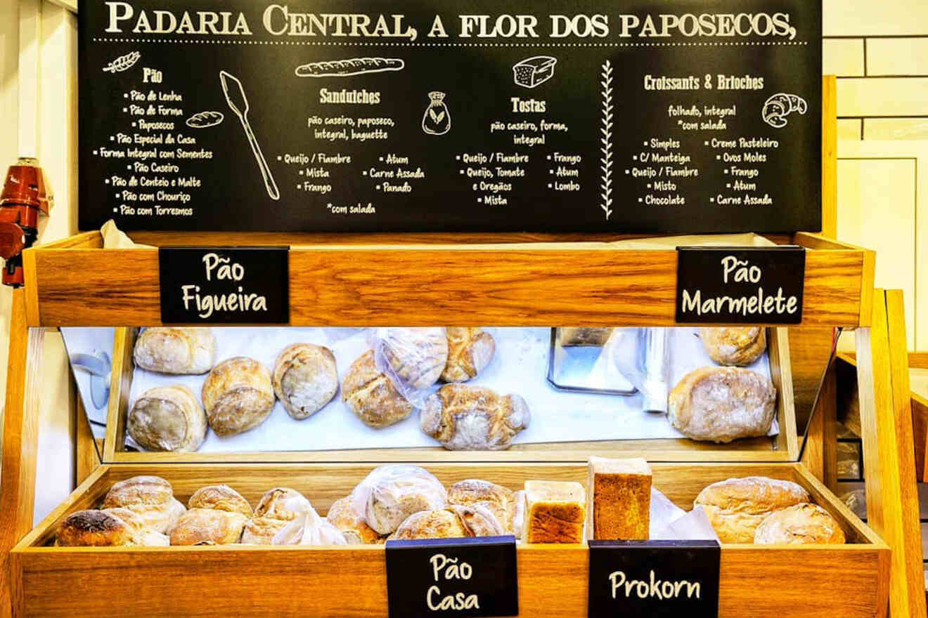 Bakery display with a variety of breads including Pão Figueira, Pão Marmilete, Pão Casa, and Prokorn. Menu board above lists sandwiches, drinks, and pastries in Portuguese.