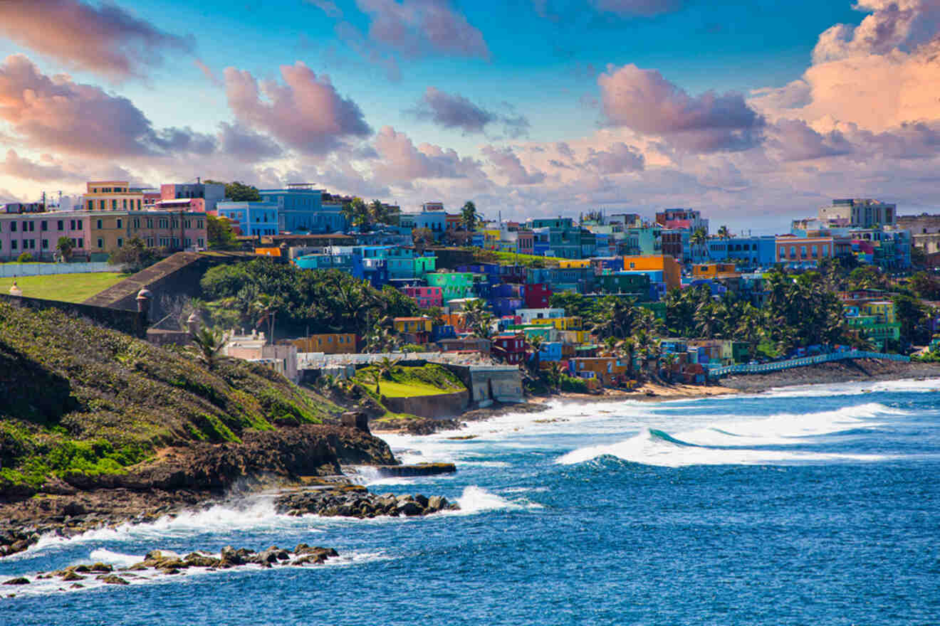 Colorful hillside buildings overlook the ocean, with waves crashing against the rocky shore and a partly cloudy sky above.