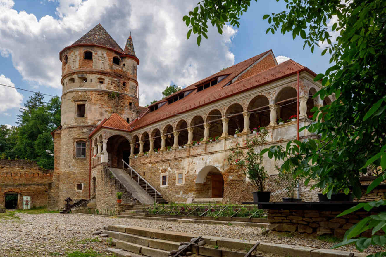 Historic stone castle with a cylindrical tower and arched walkway, surrounded by greenery and a gravel courtyard under a cloudy sky.