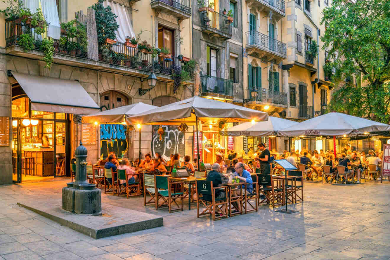 People dining outdoors under umbrellas at a bustling cafe on a charming, graffiti-adorned street corner with residential buildings in the background.