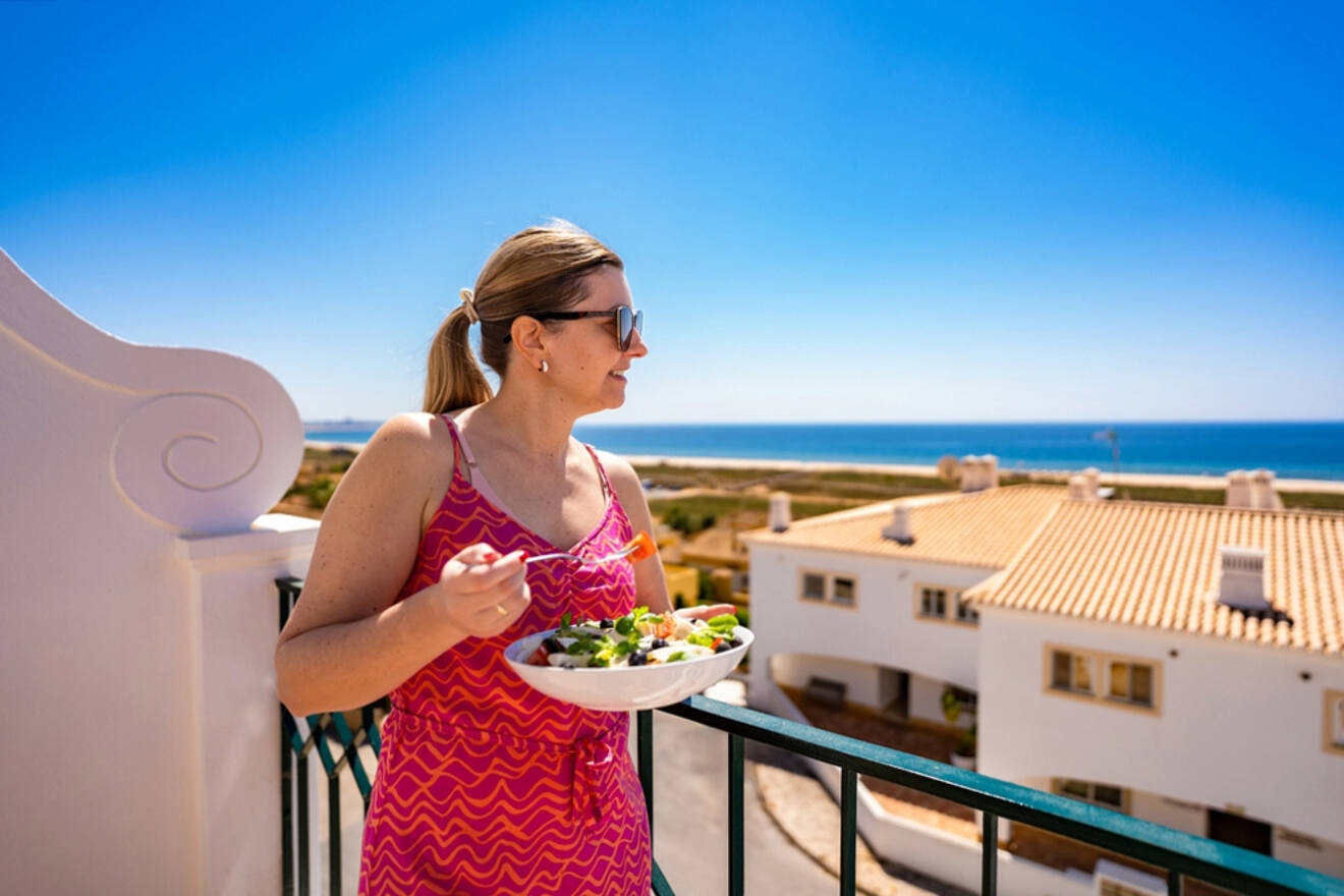 A person in a pink dress enjoys a salad on a balcony, overlooking Lagos's scenic view of white buildings and the ocean under a clear blue sky.