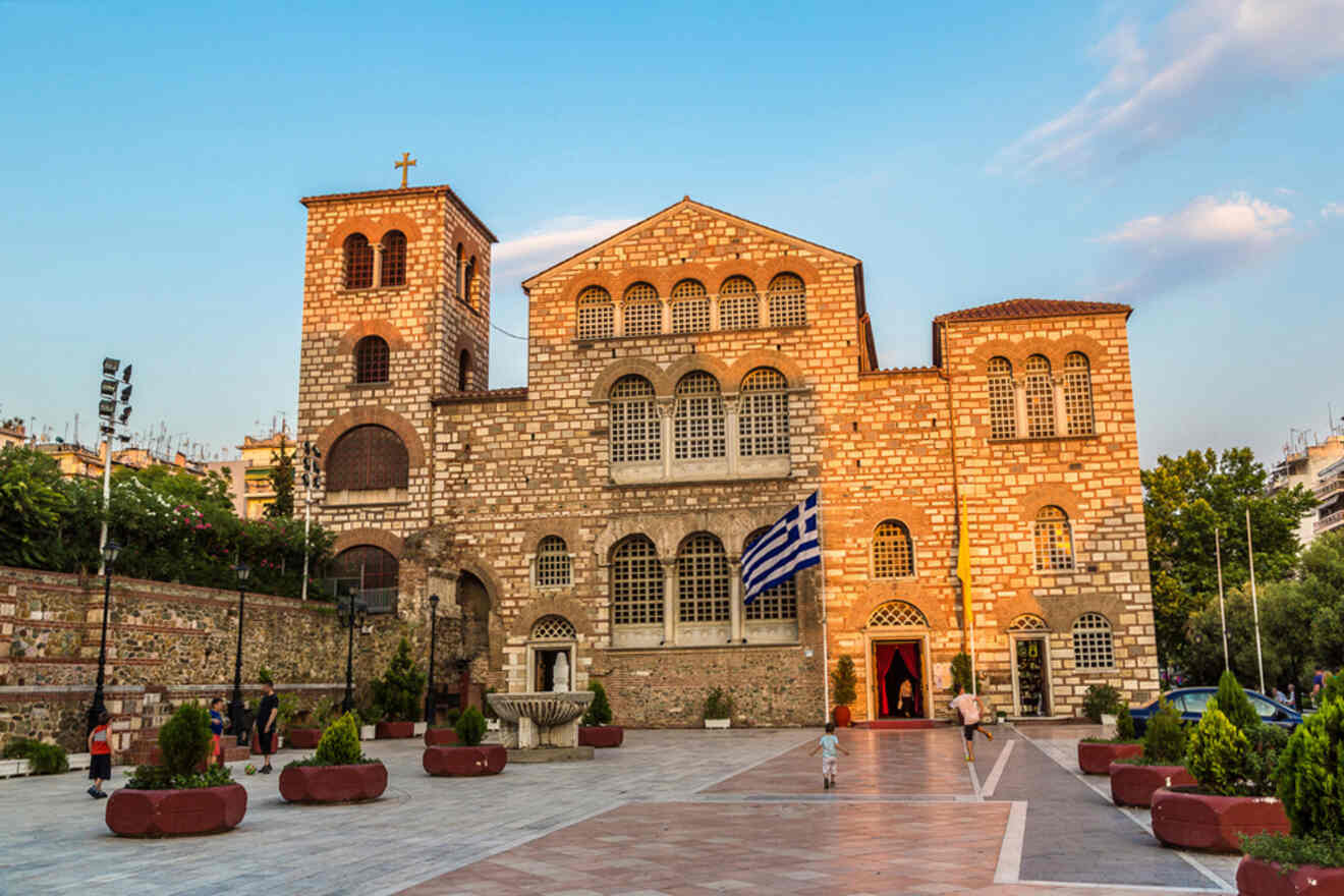 The image shows the exterior of a historic stone church with a bell tower, featuring an arched facade, a Greek flag, and a paved courtyard with potted plants.