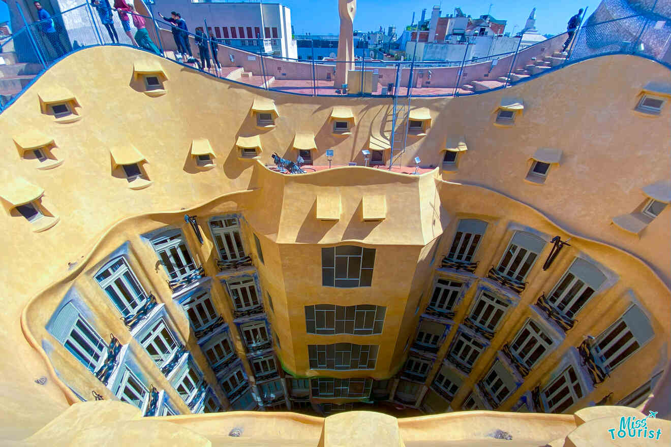 Interior courtyard of a multi-story building with a central open area surrounded by yellow walls and numerous windows. Rooftop perspective with railings visible. Blue sky above.