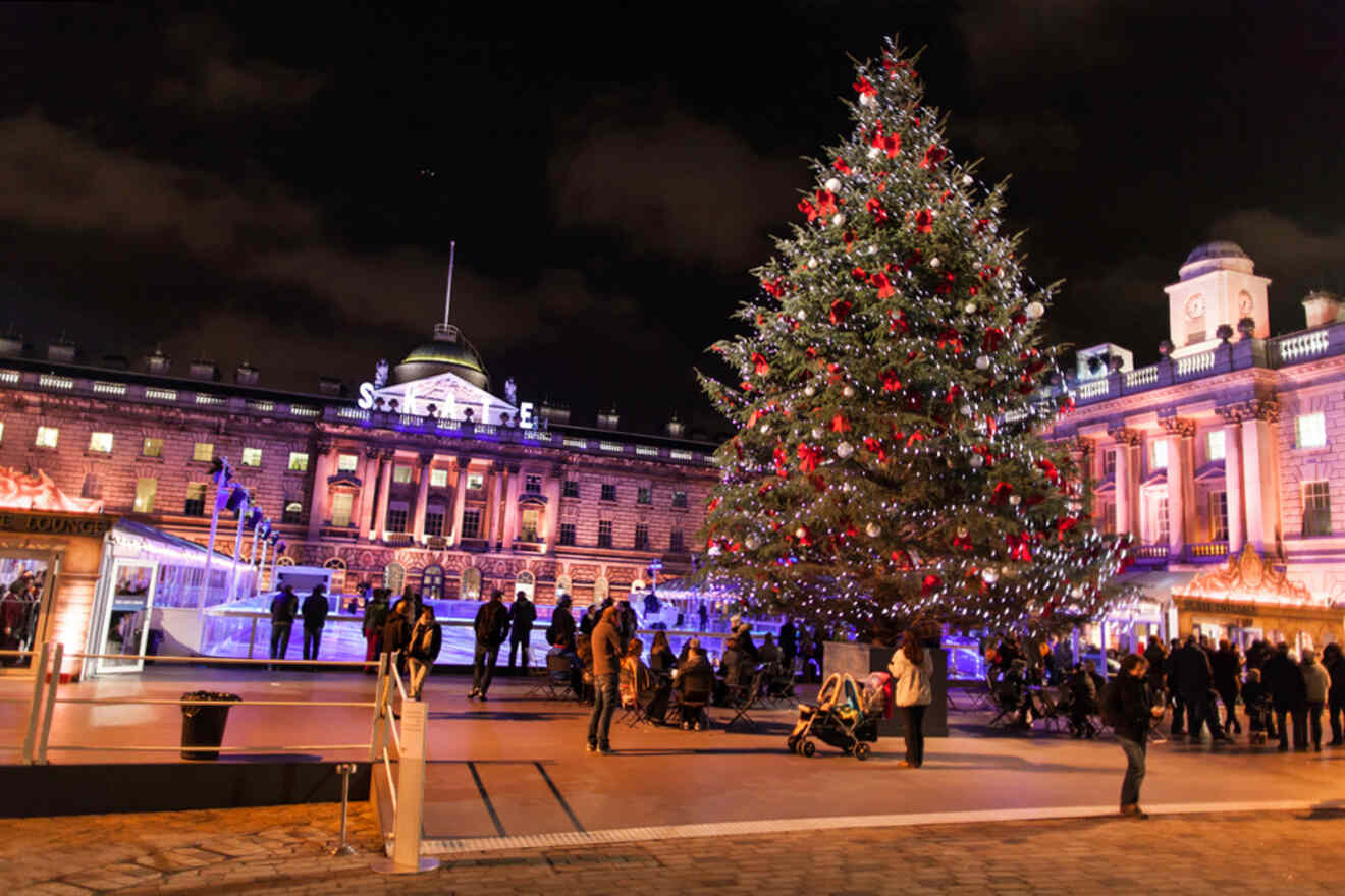 Festive scene at night with a large Christmas tree adorned with red ornaments and lights, people gathering around, and an illuminated historic building in the background.