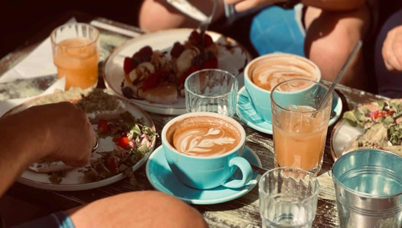 A table with cups of coffee, glasses of juice, and plates of brunch food, including waffles with toppings. People are seated around the table.